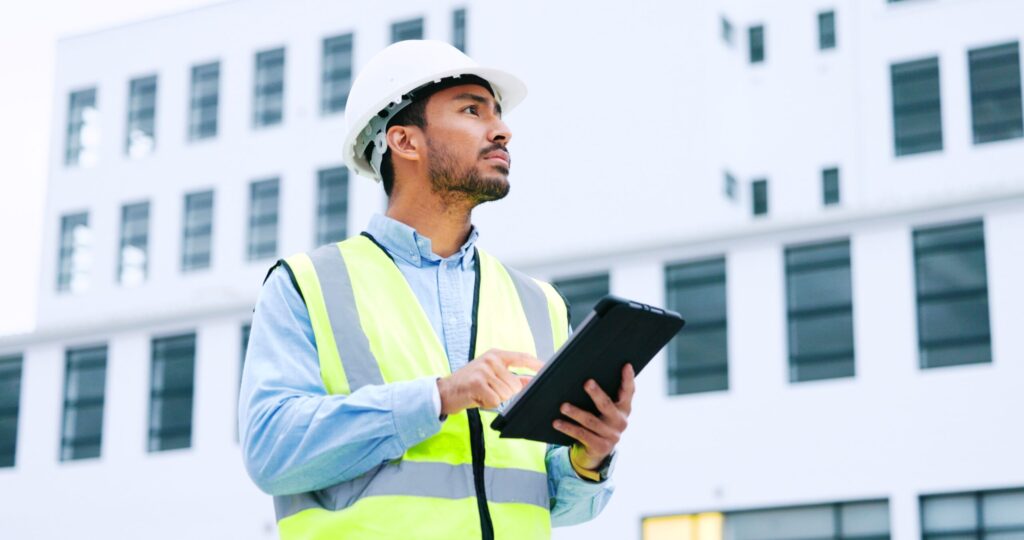 A man wearing a hard hat and high-visibility vest is looking up and to the right while holding a tablet and touching the screen with his finger. He is standing in front of a white building with many windows.